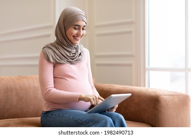 Happy Pregnant Muslim Woman Using Digital Tablet Reading Blog About Pregnancy Online Sitting On Sofa At Home. Middle Eastern Female In Hijab Browsing Internet. People And Technology Concept - Powered by Shutterstock