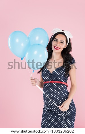 Happy pregnant woman in dotted dress holding air balloons isolated on pink