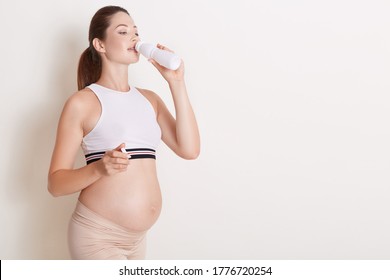 Happy Pregnant Woman Drinking Kefir Or Yogurt From Bottle, Wearing Sporty Clothes, Showing Her Bare Belly, Posing Isolated Over White Background. Copy Space.