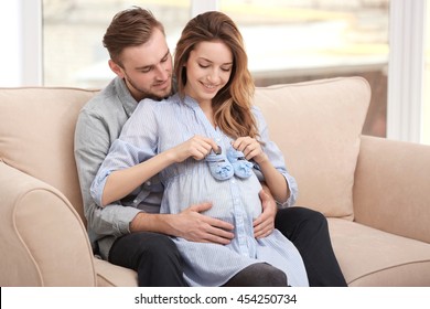 Happy pregnant woman holding baby shoes  and her husband on a sofa at home