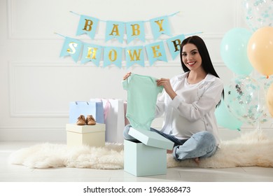 Happy Pregnant Woman Holding Onesie In Room Decorated For Baby Shower Party