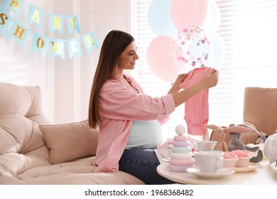 Happy Pregnant Woman Holding Onesie In Room Decorated For Baby Shower Party