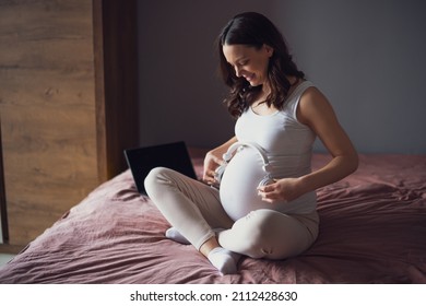 Happy Pregnant Woman Relaxing At Home. She Is Sitting On Bed In Bedroom And Playing Music To Her Baby In Stomach.