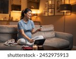 A happy pregnant woman sitting on the couch in the living room and typing in her credit card number on the tablet for online shopping.