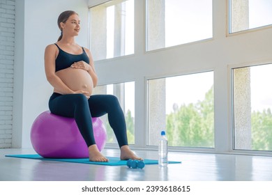 Happy pregnant woman sitting on a fitness ball and touching her belly with look in window. Caucasian expectant mother doing supportive physical exercises for smooth pregnancy