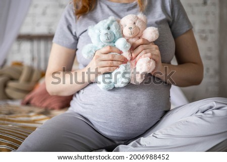 Happy pregnant woman sitting on bed at bedroom holding two cute bear toys awaiting twins baby boy and girl. Beautiful redhead female with huge tummy relaxing enjoying pregnancy waiting two at a birth