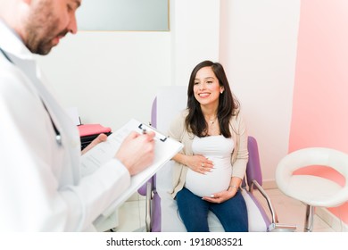 Happy pregnant woman smiling and talking with a male gynecologist about her healthy pregnancy. Doctor writing in the medical chart at the consulting room