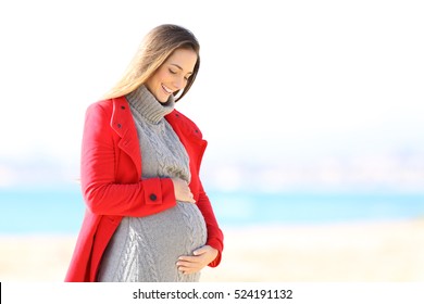 Happy pregnant woman wearing a red jacket looking her belly on the beach in winter - Powered by Shutterstock