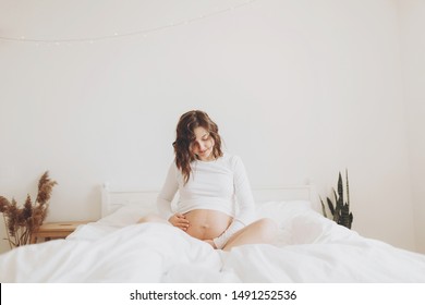 Happy Pregnant Woman In White Holding Belly Bump And Relaxing On White Bed At Home. Stylish Pregnant Mom Waiting For Baby. Motherhood And Fertility Concept. Maternity Time