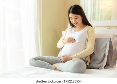 Happy pregnant young asian woman touching belly and admiring her baby or fetus on bed with happiness smile face. Young Asia mother love her baby so much. Mom waiting and expecting baby has good health - Powered by Shutterstock