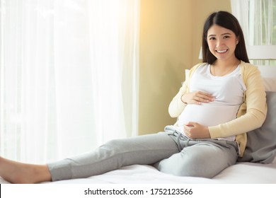 Happy pregnant young asian woman show big belly, admiring her baby or fetus on bed with happiness smile face. Young Asia mother love her baby so much. Mom expecting baby has good health smiley face - Powered by Shutterstock