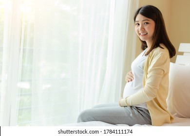 Happy pregnant young asian woman holding belly and look at camera with happiness smiley face. Young Asia pregnancy mother love her baby and has good health. Mom waiting, expecting baby has good health - Powered by Shutterstock