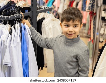 happy preschooler boy kid in clothes store is shopping, choosing. happy smile child with shirt or sport suit on hangers in hand. many, lot of different colorful clothes in shop for kids. - Powered by Shutterstock