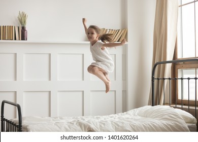 Happy Preschooler Girl Have Fun Jumping High On Comfortable Mattress On Bed At Home, Excited Little Child Entertain Enjoy Playing Alone Leap And Hop In White Bedroom Engaged In Funny Activity