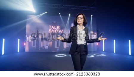 Happy presenter in a suit and glasses running onto the stage and announcing the start of the famous comedy, late-night show in an illuminated room with LED screen and 3D inscription