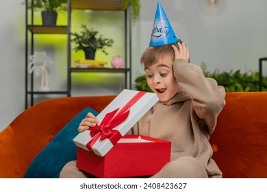 Happy preteen child boy wears festive birthday cap hat hold gift box with ribbon congratulating. Teenager male little kid celebrating party event opening delivery greeting present at home room on sofa