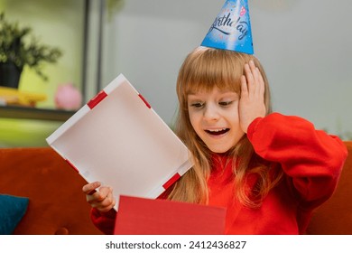 Happy preteen child girl wears festive birthday cap hat hold gift box with ribbon congratulating. Teenager amazed kid celebrating party event, opening delivery greeting present at home room on sofa