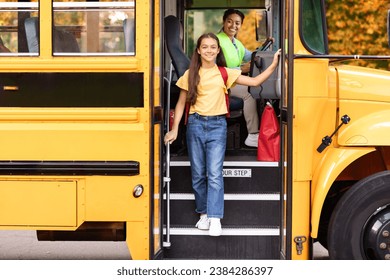 Happy preteen girl getting of the yellow school bus, cheerful female kid stepping down off of the vehicle, friendly black female driver in uniform looking at her and smiling, free space - Powered by Shutterstock
