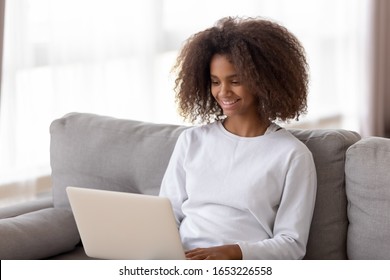 Happy Pretty African American Teen School Girl Using Laptop Device Sit On Sofa At Home, Smiling Black Teenager Student Holding Notebook Computer Surfing Social Media Chatting Online Study In Internet