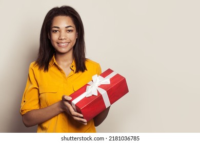 Happy pretty african american woman holding gift and looking at the camera against white wall background