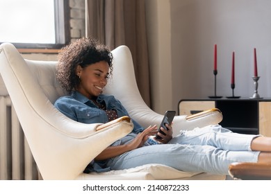 Happy pretty African girl resting in comfortable armchair, relaxing at home, using online app on mobile phone, making video call, chatting, reading text message, using virtual service on internet - Shutterstock ID 2140872233