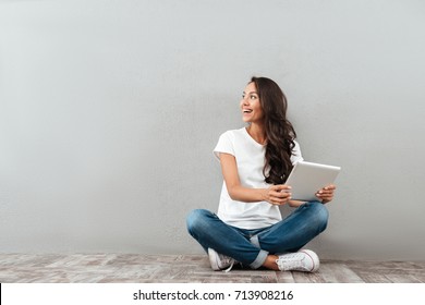 Happy hübsche Asianin, die einen Tablet-Computer hält und Kopienraum beim Sitzen einzeln auf grauem Hintergrund betrachtet