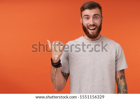 Happy pretty bearded tattooed man with trendy haircut looking at camera with wide smile and rounding his blue eyes, pointing aside with raised thumb while posing over orange background