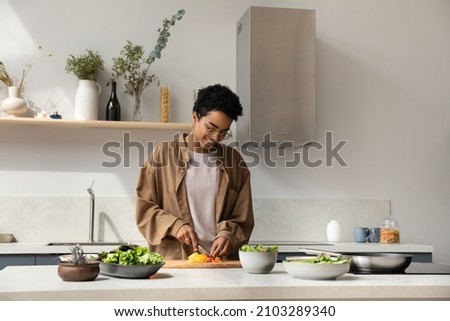 Happy pretty Black girl enjoying cooking hobby, chopping fresh vegetables for salad at kitchen table, making healthy dinner from organic bio ingredients, keeping vitamin rich nutrition