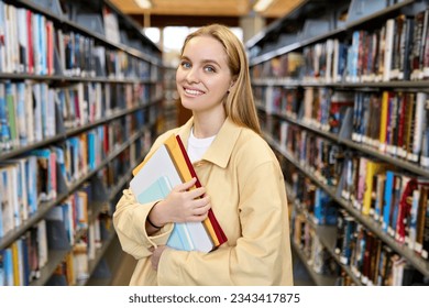 Happy pretty blonde girl, smiling female teenage high school student holding notebooks looking at camera standing in modern university or college campus library among bookshelves, portrait.
