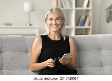 Happy pretty blonde mature woman holding credit card and mobile phone, looking at camera at home, smiling for bank customer portrait, promoting financial online service, cashback, easy payment - Powered by Shutterstock