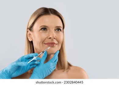 Happy pretty blonde middle aged woman gets cosmetic injection in her lips. Hyaluronic acid injection for facial rejuvenation procedure. Women in beauty salon. Plastic surgery clinic, copy space