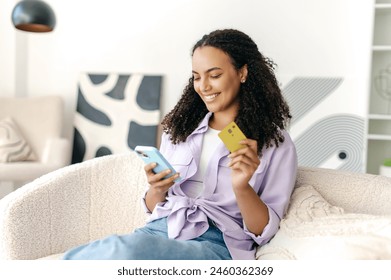 Happy pretty brazilian or hispanic curly haired woman, holding smart phone and credit card for payment, online shopping or transaction, ordering delivery while sitting in living room at home, smiling - Powered by Shutterstock