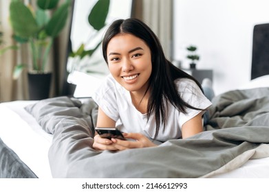 Happy Pretty Chinese Girl Using Her Smart Phone While Lying On A Bed At Home, Texting On Cell Phone, Reading News Online, Browsing Social Media, Looks At Camera Smiling. Gadgets And Technology Concept