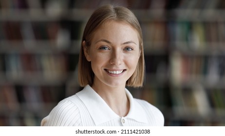Happy pretty college student girl posing in university library with blurred bookshelves behind. Young millennial woman looking at camera with toothy smile head shot portrait