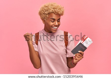 Happy pretty dark-skinned woman holds passport and plane tickets, rejoices to have tour abroad with boyfriend, has special offer from travel company, isolated over pink background. Cheap Flight Deals.