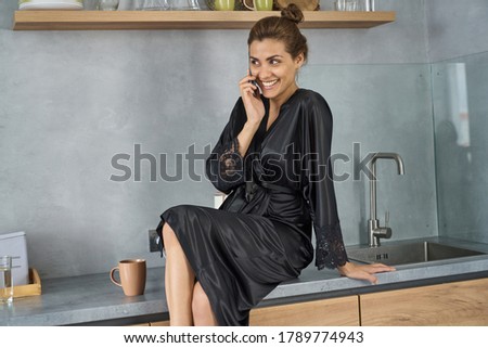 Happy pretty female in black silk dressing gown is sitting on table and enjoying phone conversation while drinking coffee