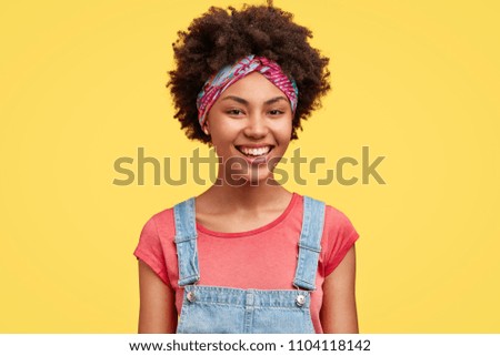 Happy pretty female housewife dressed in casual t shirt and denim overalls, has broad smile as did all work about house, poses against yellow background indoor. African American woman giggles