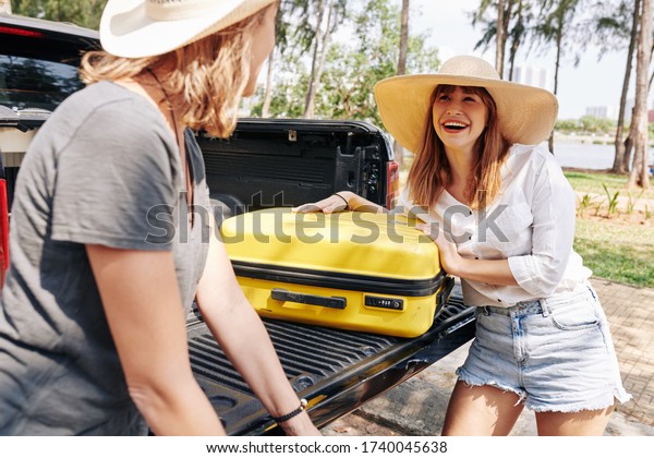 Happy pretty friends in straw hats
taking suitcases out of car trunk after coming to
beach