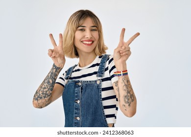 Happy pretty gen z blonde young woman, smiling girl with short blond hair tattoos wearing striped t-shirt denim dress rainbow bracelet showing peace sign standing isolated on white. Portrait. - Powered by Shutterstock