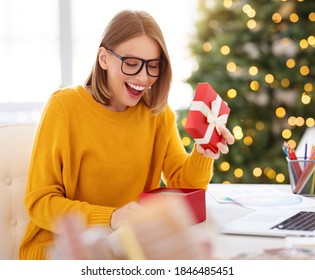 Happy Pretty Girl Freelancer Smiling And Opens A Holiday Gift During Remote Work At Laptop At Home  Office Before Christmas
