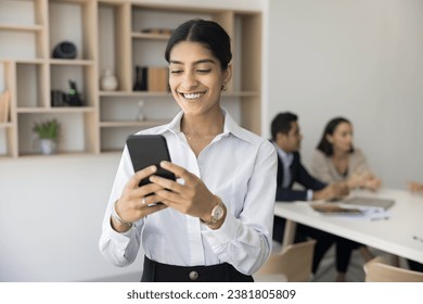 Happy pretty Indian manager girl using mobile phone for work communication, typing, chatting, making call prom office meeting room, co-working space with colleagues talking behind - Powered by Shutterstock