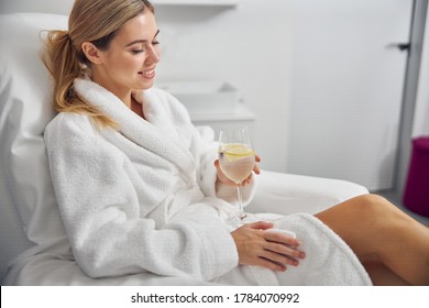 Happy pretty lady is sitting in bathrobe in pedicure chair and enjoying glass of chilling water with lemon - Powered by Shutterstock