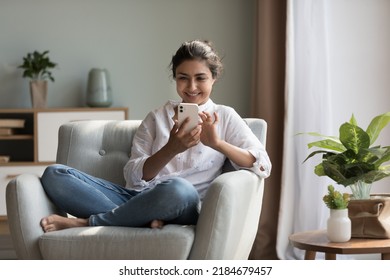 Happy pretty millennial Indian girl relaxing at home, resting in armchair, typing on smartphone, using online app, software, shopping on Internet, making video call. Mobile phone communication - Shutterstock ID 2184679457