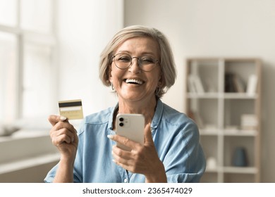 Happy pretty older retired bank customer promoting credit card, using financial e-commerce application, service on smartphone, paying for purchase on Internet, looking at camera for portrait - Powered by Shutterstock