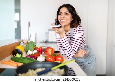 Happy pretty short-haired brunette young middle eastern woman recording voice message using smartphone while cooking healthy organic food at home, looking at copy space and smiling, kitchen interior - Powered by Shutterstock