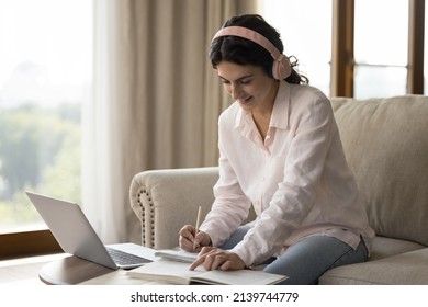 Happy Pretty Student Girl In Trendy Big Headphones Watching Virtual Class On Laptop, Listening To Audio Lecture, Reading Book, Writing Notes, Practicing Multitasking, Preparing For College Test