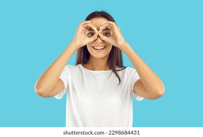 Happy pretty woman looking through pretend binoculars. Cheerful beautiful young brunette lady in white T shirt forming circles with her hands, doing binoculars gesture, looking in distance and smiling - Shutterstock ID 2298684431