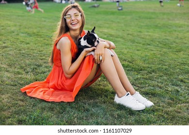 Happy Pretty Woman Sitting On Grass In Summer Park, Holding Boston Terrier Dog, Smiling Positive Mood, Wearing Orange Dress, Trendy Style, Slim Legs, Sneakers, Playing With Pet