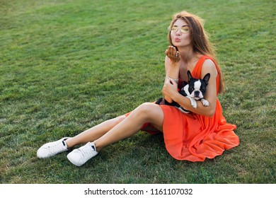 Happy Pretty Woman Sitting On Grass In Summer Park, Holding Boston Terrier Dog, Kiss, Wearing Orange Dress, Trendy Style, Slim Legs, Sneakers, Playing With Pet