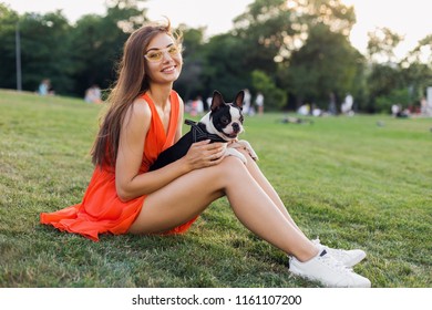 Happy Pretty Woman Sitting On Grass In Summer Park, Holding Boston Terrier Dog, Smiling Positive Mood, Wearing Orange Dress, Trendy Style, Slim Legs, Sneakers, Playing With Pet